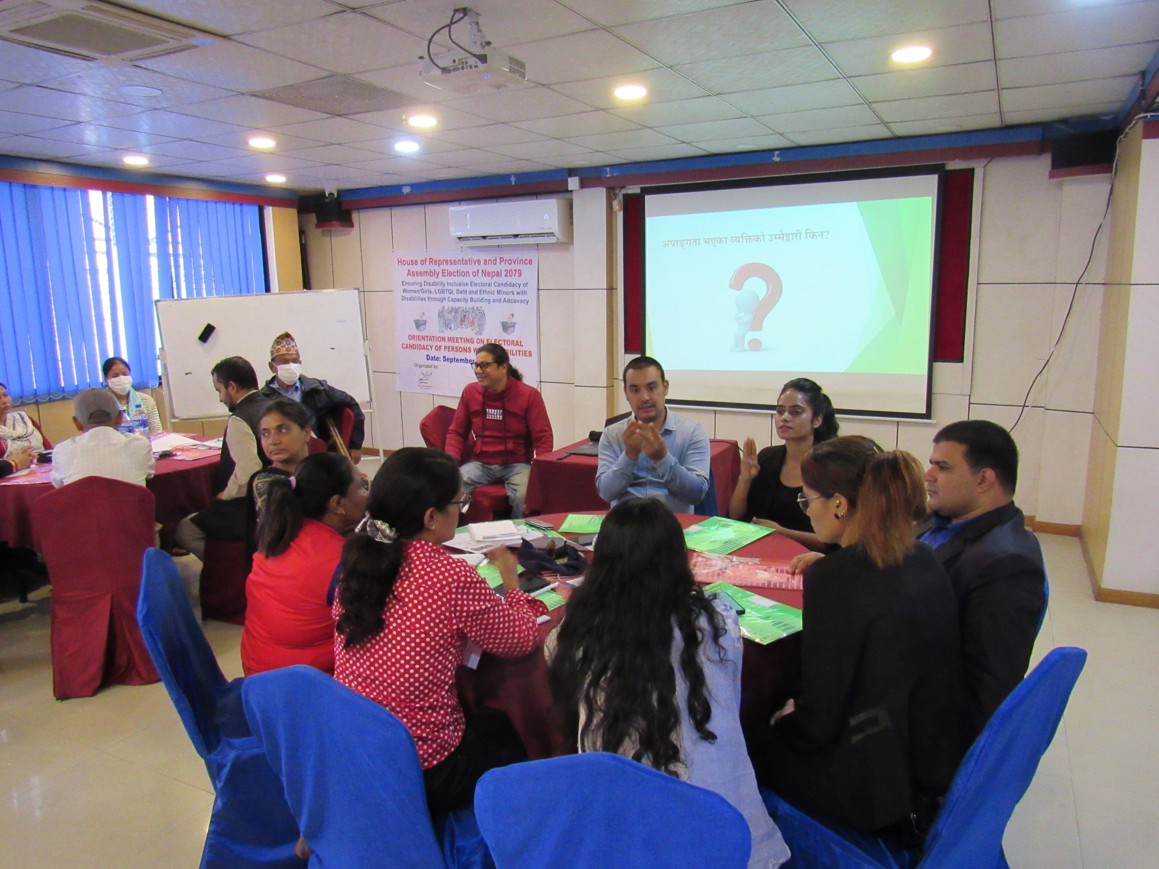 around 25 People are being seated in the round table setup and facilitator is presenting in the front